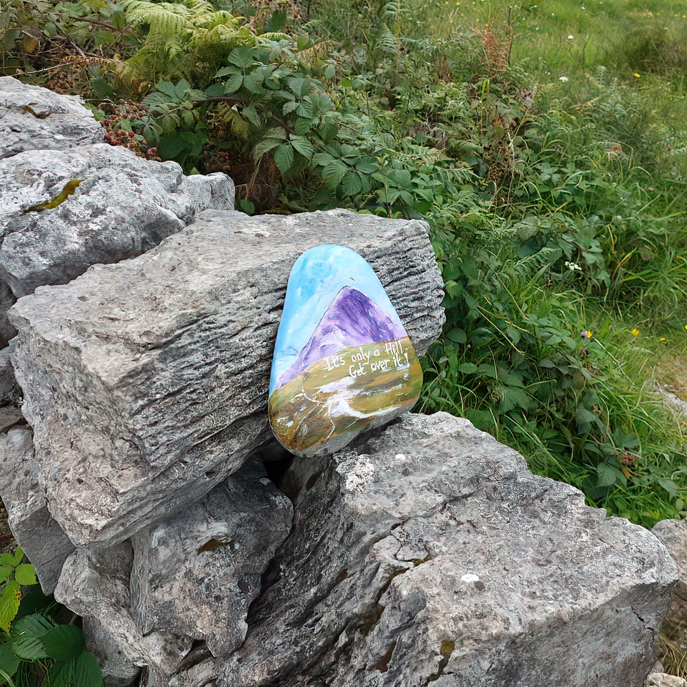 A colourfully painted rock with the motivational message "It's only a Hill. Get over it!" painted on, balanced on a small stone wall